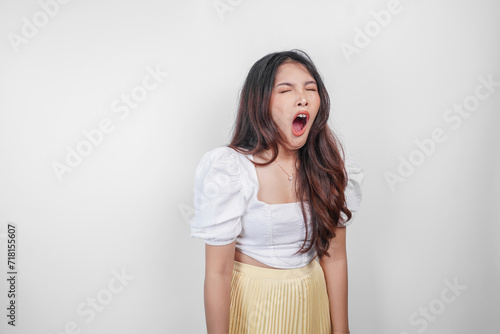 Portrait of sleepy attractive Asian woman wearing a dress, feeling tired after night without sleep, yawning with her mouth wide open photo