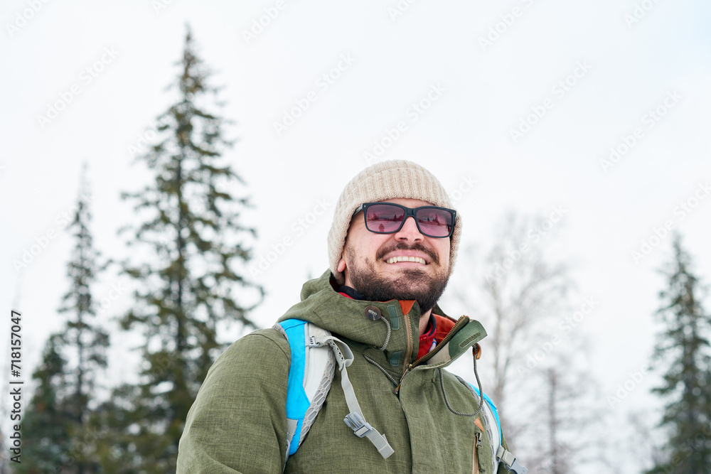 Fototapeta premium Cheerful handsome bearded backpacker in sunglasses looking into distance enjoying beauty of winter landscape