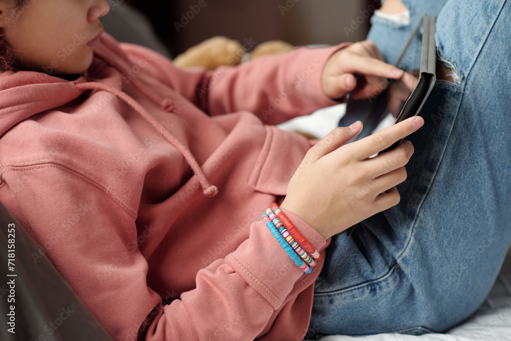 Hand of female teenage schoolgirl in pink hoodie and blue jeans holding tablet while sitting in front of camera and working over project