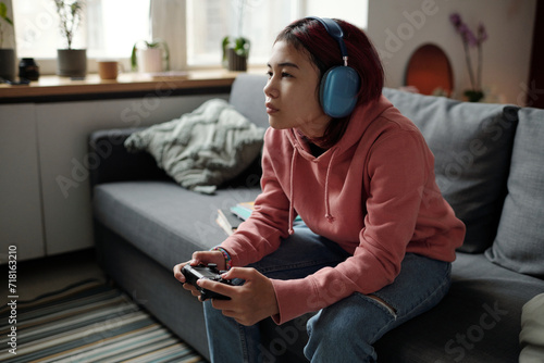 Serious teenage girl in headphones sitting on couch in front of camera and pressing buttons of joystick while playing video game