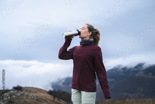 A young cute girl with curly brown ponytail hairstyle drinks water from a thermal glass in the mountains, a tourist drinks a hot drink at a rest stop, above the clouds in the mountains in hiking cloth