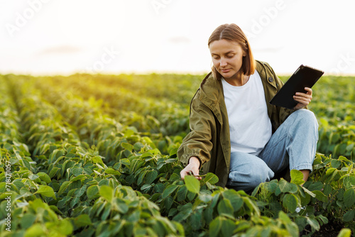 Modern agribusiness. Female farmer with digital tablet examines and checkins green leaves of soybeans plants at field. Agronomist controls growth and development of sprouts. Smart farming technology.