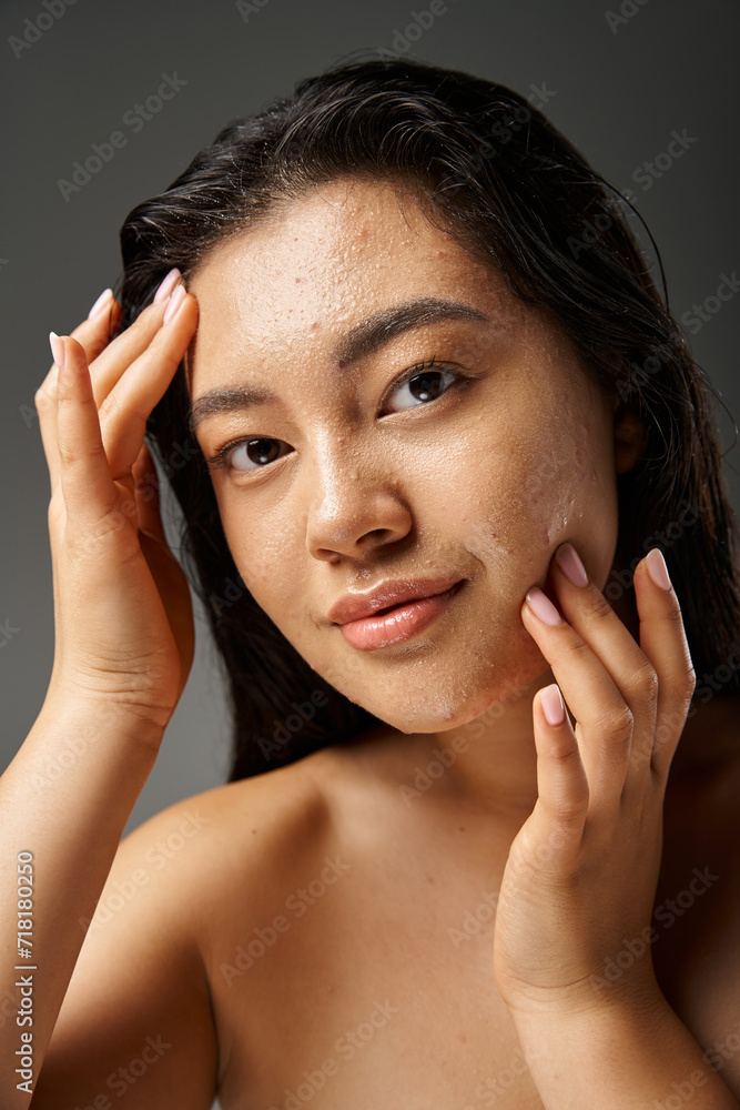 young asian woman with brunette hair toughing wet face with acne and looking at camera on grey young asian woman with brunette hair toughing wet face with acne and looking at camera on grey