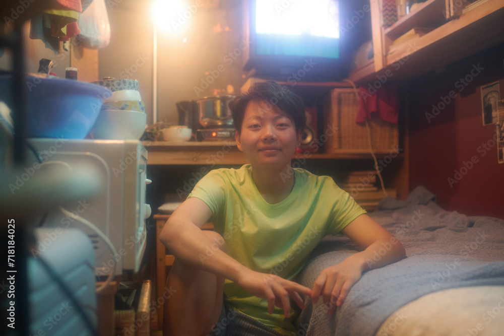 Happy Asian girl sitting on the floor by single bed in small apartment and looking at camera against wooden table with kitchenware