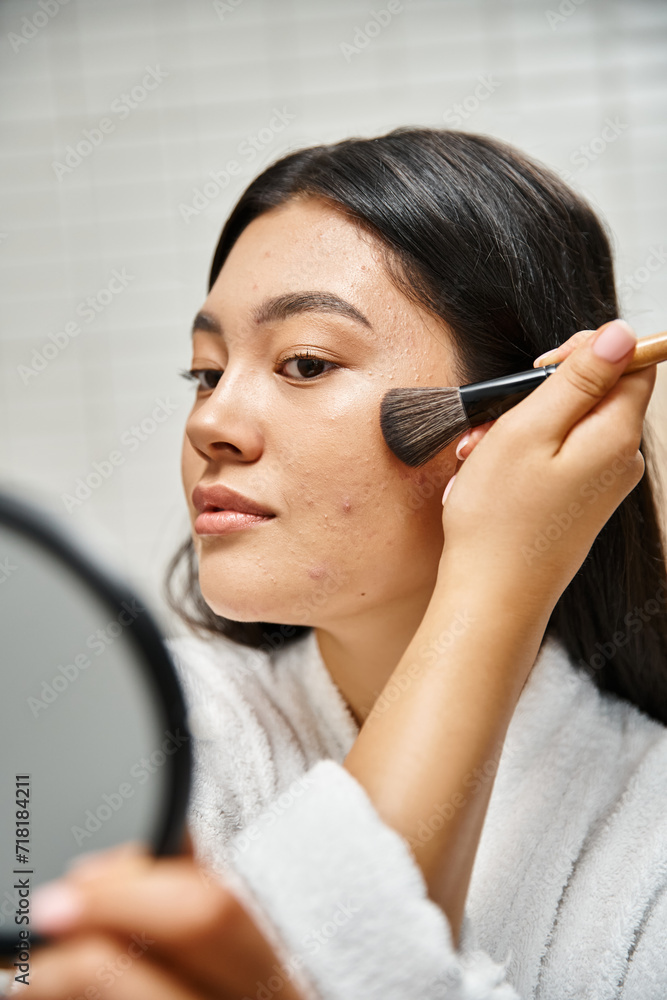 brunette young asian woman applying face powder over acne-prone skin with cosmetic brush, makeup