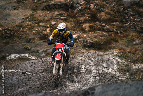 Portrait of professional motorcyclist in full moto equipment riding crops enduro bike in mountains, driver rides and dirt and stones fly off the wheel, motorcycle race, concept of motosport