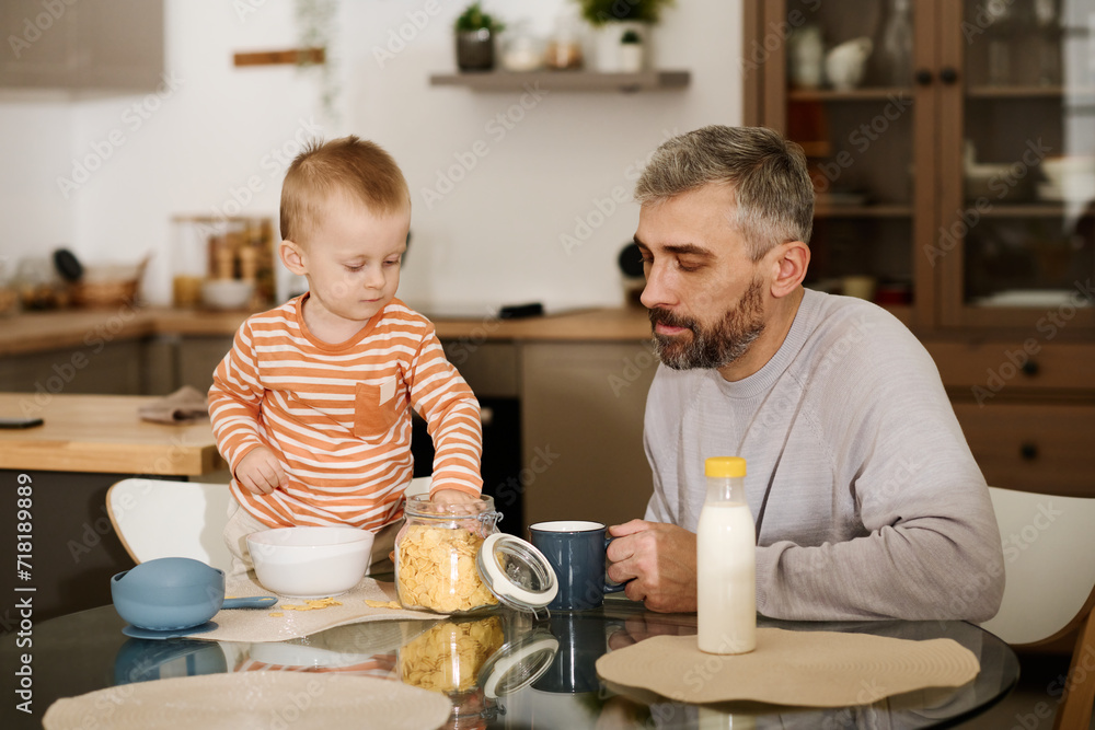 Mature father sitting by kitchen table and looking at his adorable baby son taking some cornflakes out of open jar during breakfast