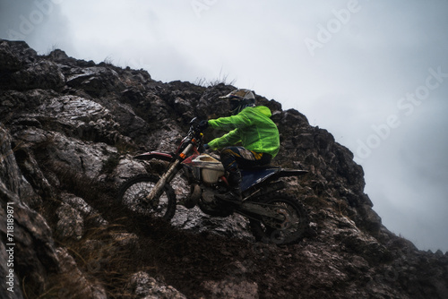A racer at a competition rides along a cliff in rainy and gloomy weather, bottom view, professional motorcyclist in full moto equipment riding crops enduro bike in highlands photo