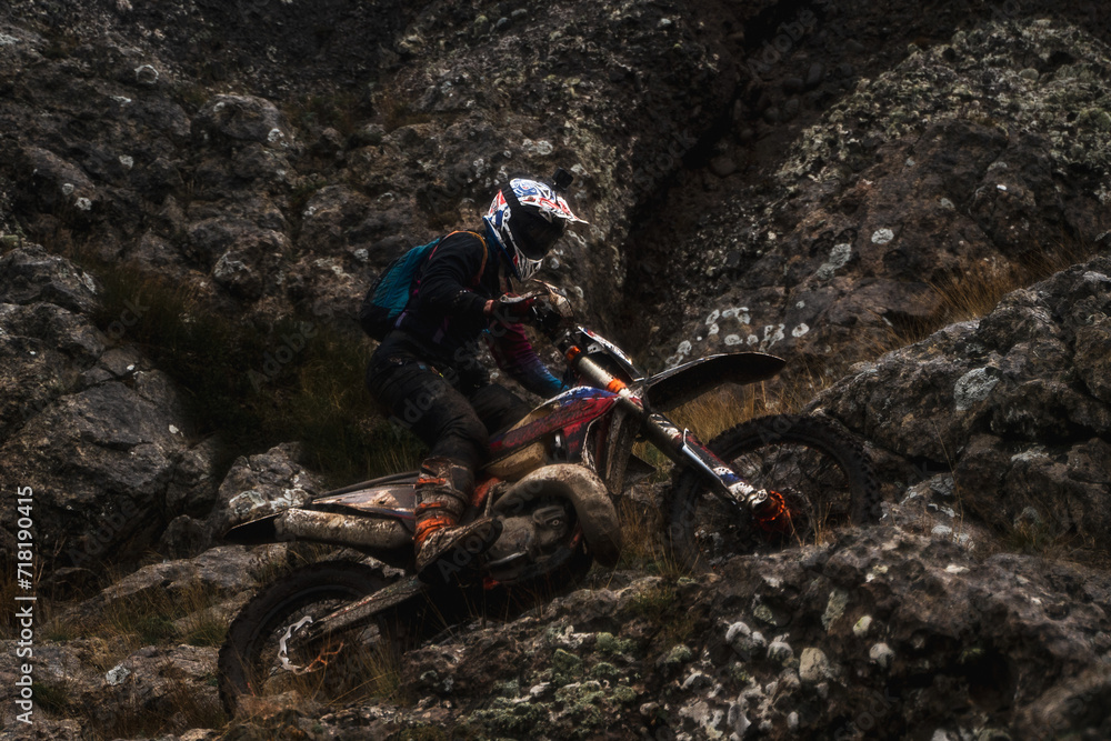 Obraz premium A racer at a competition rides along a cliff in rainy weather, bottom view, professional motorcyclist in full moto equipment riding crops enduro bike in highlands, concept of motosport