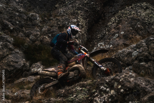 A racer at a competition rides along a cliff in rainy weather, bottom view, professional motorcyclist in full moto equipment riding crops enduro bike in highlands, concept of motosport
