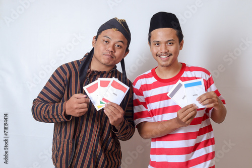 Portrait of two excited Indonesian man from Javanese and Madurese tribes holding voting paper for General Election (Pemilu) of president and government of Indonesia. Isolated image on gray background photo