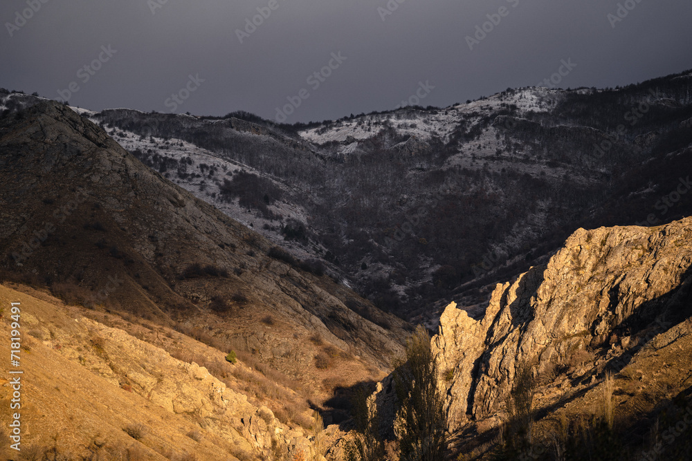 Dramatic landscape of rocks at golden hour, sunrise in snowy mountains, gloomy gray sky, snowy forest and yellow sun light