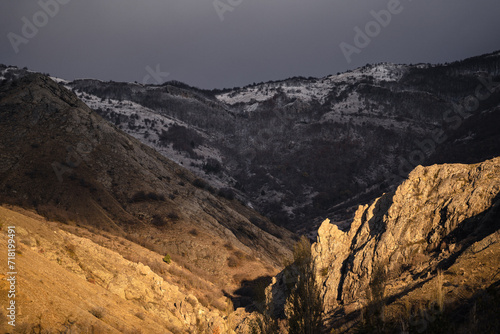Dramatic landscape of rocks at golden hour, sunrise in snowy mountains, gloomy gray sky, snowy forest and yellow sun light
