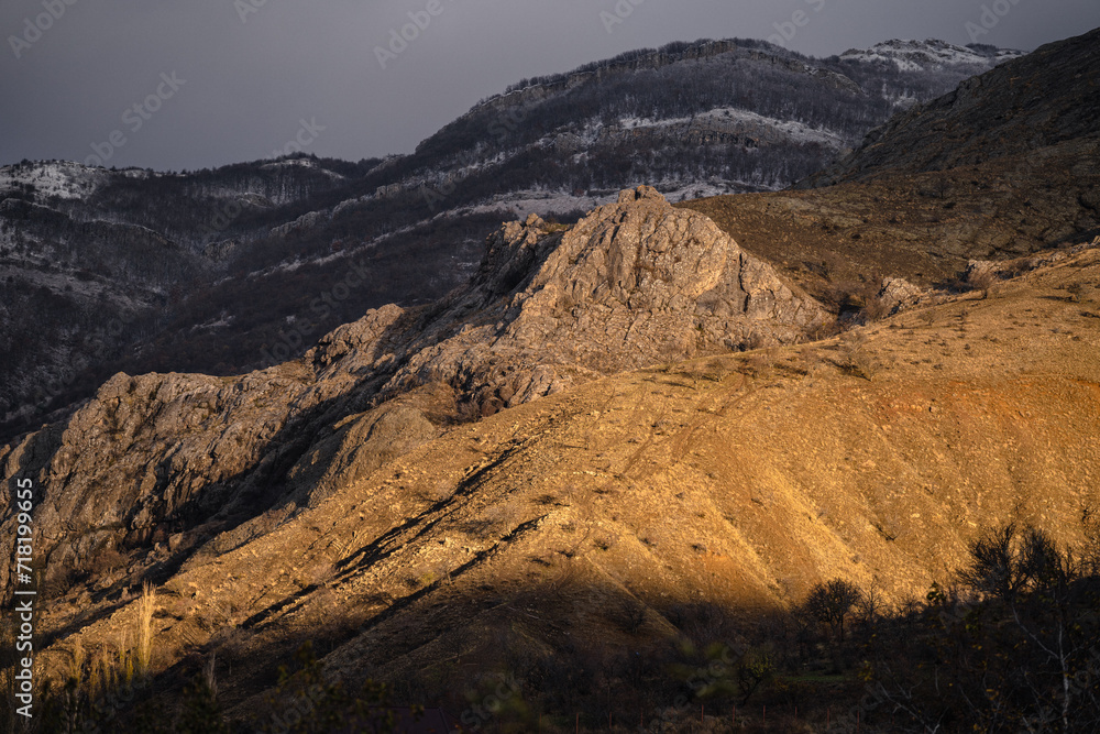 Fototapeta premium Landscape of rocks at classic golden hour glow, sunrise in snowy mountains, dramatic gray sky, snowy forest and yellow sun light, layers of mountains