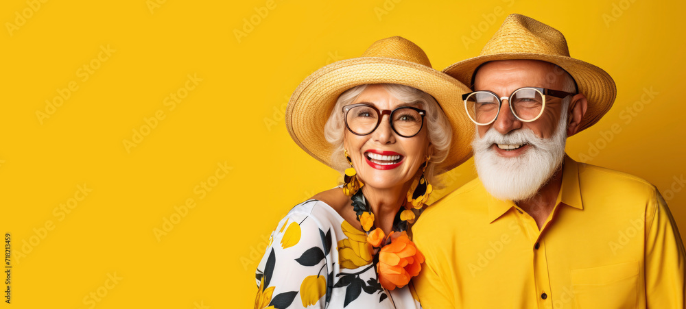 Studio portrait of stylish cheerful loving pensioners in bright summer clothes on yellow background. Happy mature man and woman in straw hats ready for vacation. Summer holiday and travel concept