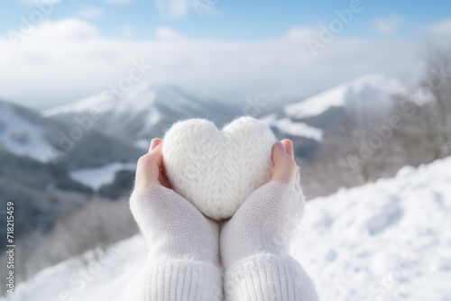  a person's hands holding a white heart shaped object in front of a snowy mountainside with a mountain range in the backgrouch of the picture.