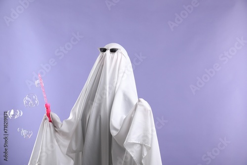 Funny ghost. Person in white sheet, sunglasses and soap bubbles on violet background, space for text