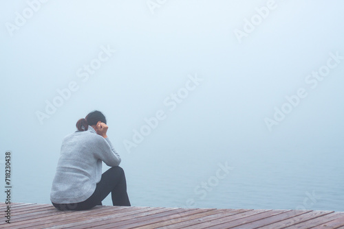 Junge Frau sitzt einsam am Rand eines Holzweges einer Brücke gebückt und traurig in Gedanken versunken im Nebel photo