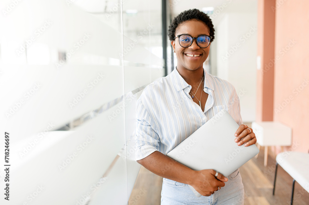 Purposeful African-American businesswoman in smart casual wear stand in modern coworking space and holding laptop, female freelancer or student with laptop computer indoors and looking at camera Purposeful African-American businesswoman in smart casual wear stand in modern coworking space and holding laptop, female freelancer or student with laptop computer indoors and looking at camera