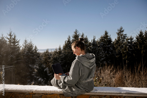 Frau im Bademantel sitzt auf einer verschneiten Steinmauer in der Natur und arbeitet am laptop