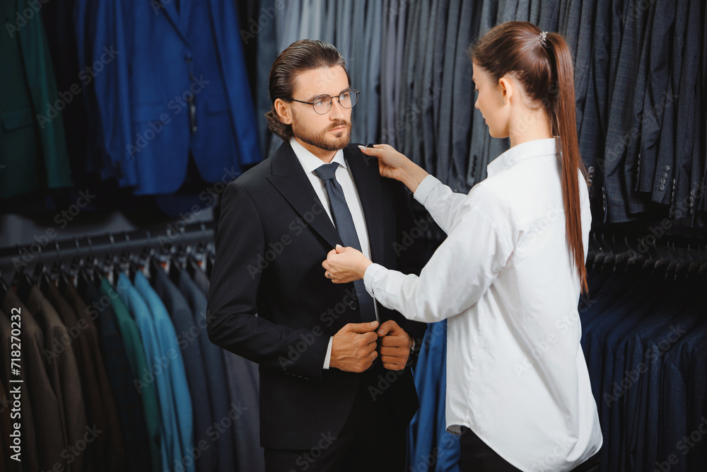 Beautiful woman seller smiling and help with choice suit to modern young handsome businessman in shop of classic clothes for men