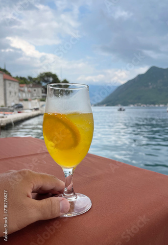 Hand holds a glass of juice against a background of sea and mountains while traveling in Europe