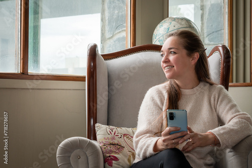 Mujer mirando por la ventana sonriente mientas usa su celular, sentada cómodamente en su sillón photo