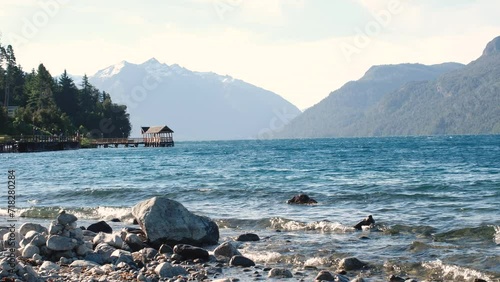 Tranquil Beauty in 4k. A Serene Rocky Beach with Majestic Mountains, Lush Trees, and a Pier Jutting into Crystal Clear Waters. High Resolution.
