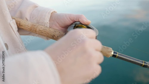 Woman hobby fishing on sea tightens fishing line reel of fish summer. Handle rotation with reel of fishing rod against of orange sunset slow motion. Calm surface sea. Hobby concept photo