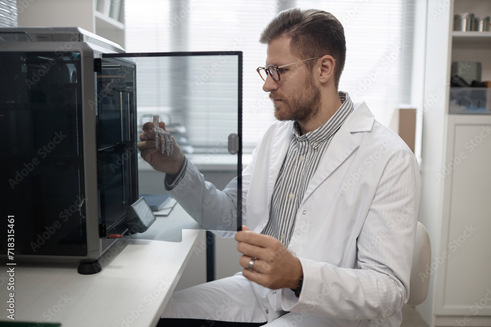 Medium long shot of Caucasian man engineer in lab coat and eyeglasses taking produced bone sample out of enclosed 3D printer in laboratory
