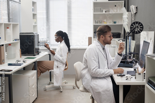Side view of Caucasian male technician holding 3D dental model and looking at computer screen while sitting at desk in laboratory  Black female colleague working in blurred background