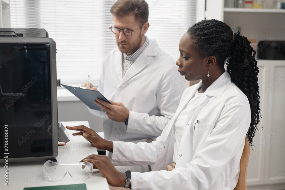 African American woman technician activating settings on touchscreen controller of enclosed 3D Printer while sitting at laboratory desk with defocused male coworker writing in clipboard African American woman technician activating settings on touchscreen controller of enclosed 3D Printer while sitting at laboratory desk with defocused male coworker writing in clipboard