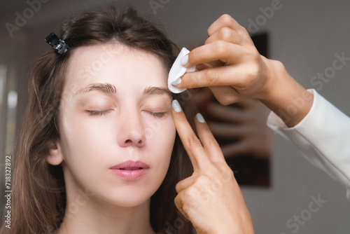 Quadro su tela In a beauty salon, a master cosmetologist applies makeup and wipes eyebrows with a cotton pad