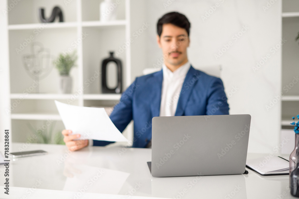 A professional Hispanic businessman in a blue blazer is focused on reading a document while sitting at a modern office desk, his laptop open in front of him. Male employee preparing annual report A professional Hispanic businessman in a blue blazer is focused on reading a document while sitting at a modern office desk, his laptop open in front of him. Male employee preparing annual report