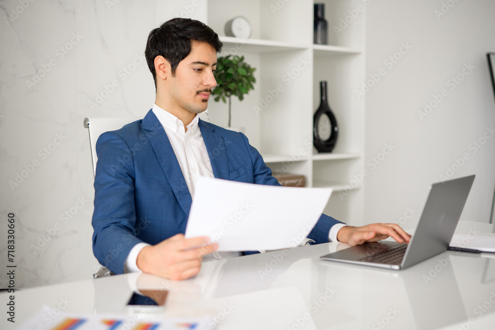 A thoughtful Hispanic businessman, wearing a blue jacket is reviewing a document while multitasking on his laptop in a contemporary office in a moment of critical thinking and business analysis A thoughtful Hispanic businessman, wearing a blue jacket is reviewing a document while multitasking on his laptop in a contemporary office in a moment of critical thinking and business analysis