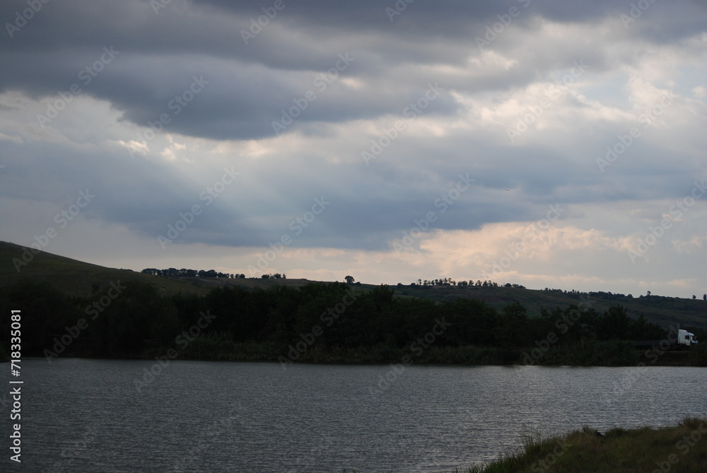 Wild steppe nature. Trees, bushes, grass with green foliage grow on a hillside under a sky with gray clouds. A small pond with light gray water at the bottom. The sun's rays break through the clouds. Wild steppe nature. Trees, bushes, grass with green foliage grow on a hillside under a sky with gray clouds. A small pond with light gray water at the bottom. The sun's rays break through the clouds.