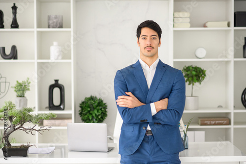 A confident Hispanic businessman stands with crossed arms in a modern office, his poised stance complementing the sleek, contemporary office. Professionalism and determination concept