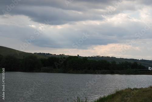 Wild steppe nature. Trees, bushes, grass with green foliage grow on a hillside under a sky with gray clouds. A small pond with light gray water at the bottom. The sun's rays break through the clouds.