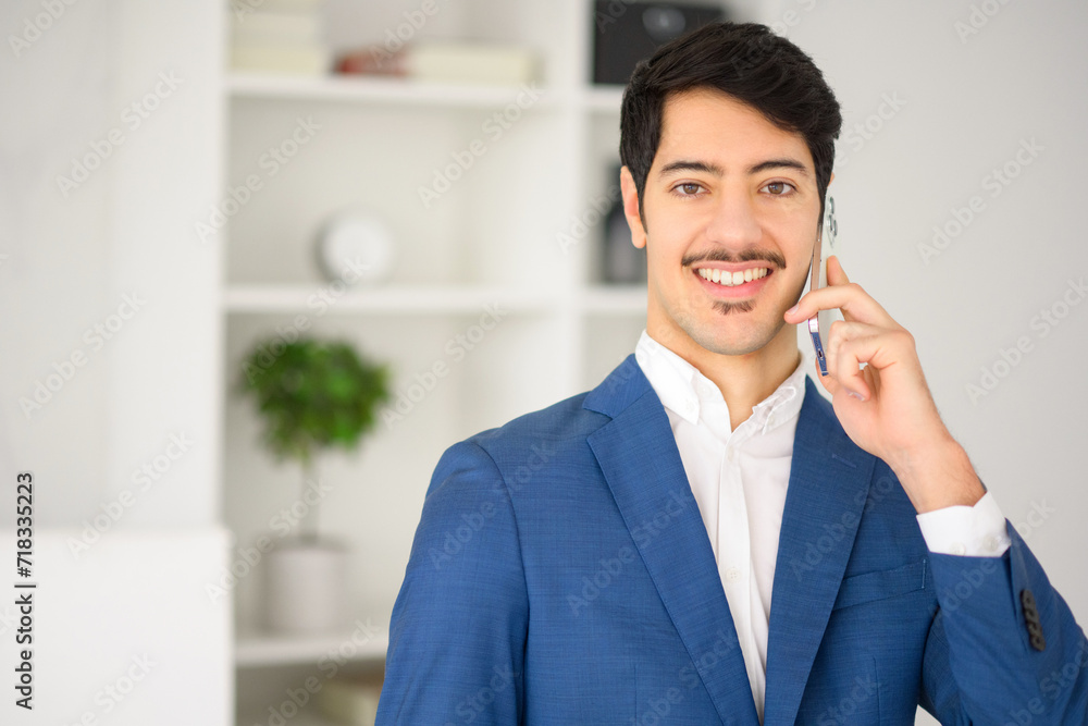 Charismatic Hispanic businessman in blue suit is engaging in conversation over smartphone, set against modern office, signaling blend of traditional business conduct and contemporary workspace design Charismatic Hispanic businessman in blue suit is engaging in conversation over smartphone, set against modern office, signaling blend of traditional business conduct and contemporary workspace design