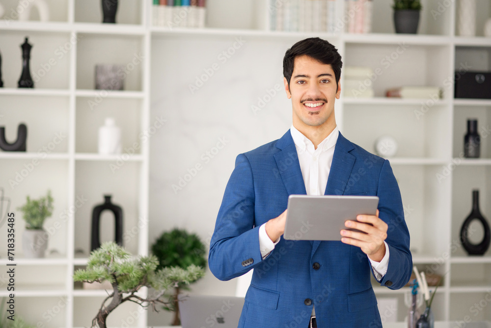 In a modern office setting, a Hispanic businessman expertly navigates his tablet, looking at the camera with a bright smile standing in modern office In a modern office setting, a Hispanic businessman expertly navigates his tablet, looking at the camera with a bright smile standing in modern office