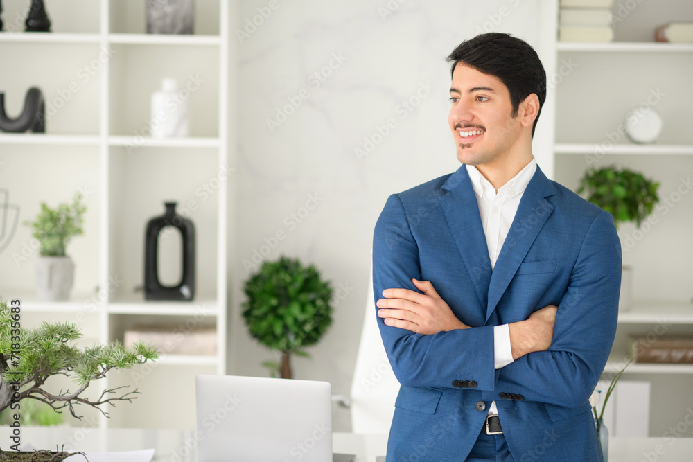 Young businessman smiling and standing in a relaxed pose with arms crossed in a bright office, communicate friendliness and a welcoming business atmosphere. A positive work environment concept Young businessman smiling and standing in a relaxed pose with arms crossed in a bright office, communicate friendliness and a welcoming business atmosphere. A positive work environment concept