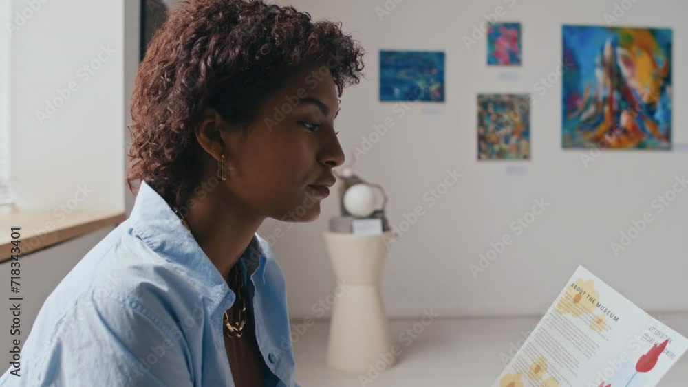 Medium close-up tilting shot of hands and face of young curly African American woman sitting on couch in art gallery at modern exhibition and reading information leaflet Medium close-up tilting shot of hands and face of young curly African American woman sitting on couch in art gallery at modern exhibition and reading information leaflet