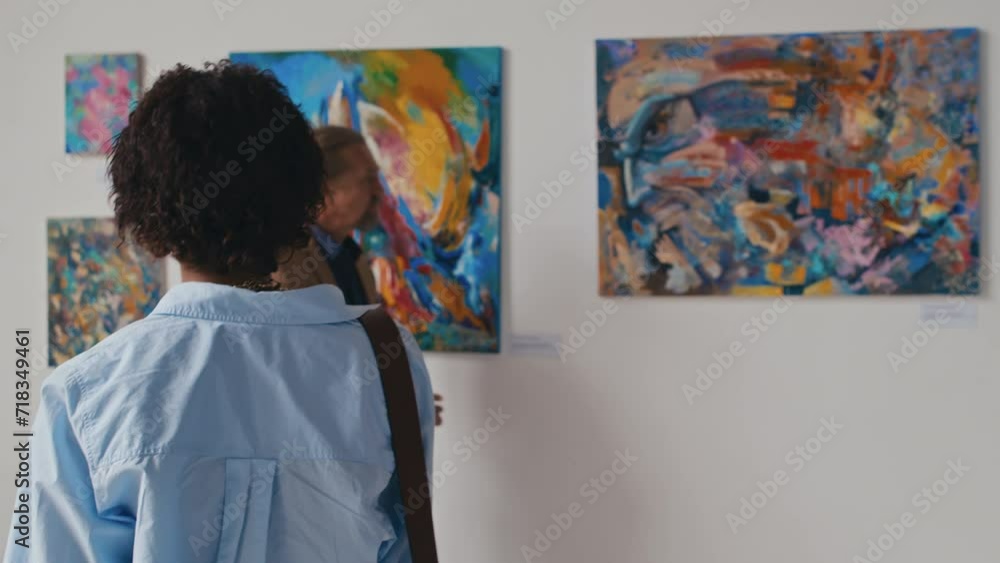 Medium close-up follow shot of young casually dressed African American woman walking through museum hall towards wall with colourful abstract paintings, stopping and viewing them closely Medium close-up follow shot of young casually dressed African American woman walking through museum hall towards wall with colourful abstract paintings, stopping and viewing them closely