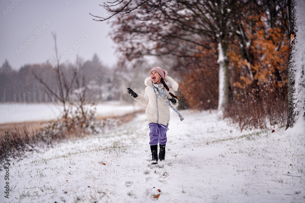 a small child in winter clothing joyfully running on a snowy path lined with bare trees and autumn leaves