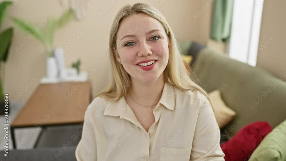 A cheerful young woman with blonde hair and a beige blouse sits in a modern living room with a sofa and houseplants. A cheerful young woman with blonde hair and a beige blouse sits in a modern living room with a sofa and houseplants.