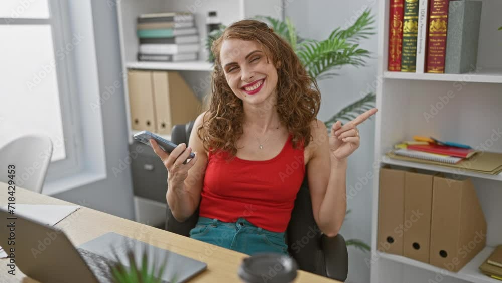 Cheerful young woman pointing to the side, using her smartphone with a joyous smile at the office Cheerful young woman pointing to the side, using her smartphone with a joyous smile at the office