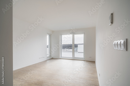 Unfurnished living room in new apartment. The room has large window and wooden floor. Walls are bright white and are equipped with electrical outlets,controls and other connections. Shades are opened.