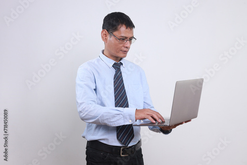 Confident adult Asian man in shirt and tie working on laptop while standing against white background