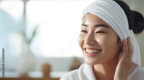 A Closeup head shot of a young beautiful Asian woman applying facial moisturizer after bathing. Smiling beautiful woman wrapped in towel to smooth her skin Daily routine in the morning. photo