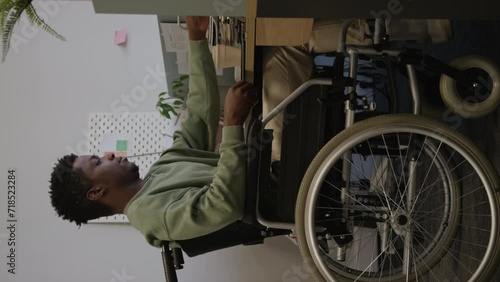 Vertical side view full shot of young African American wheelchaired male employee using laptop computer at desk in modern workspace photo